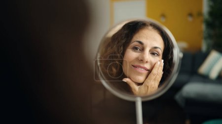 Foto de Middle age hispanic woman sitting on sofa looking face on mirror at home - Imagen libre de derechos