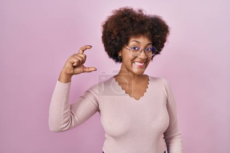 Téléchargez les photos : Jeune femme afro-américaine debout sur fond rose geste souriant et confiant avec la main faisant signe de petite taille avec les doigts à la recherche et la caméra. concept de mesure. - en image libre de droit