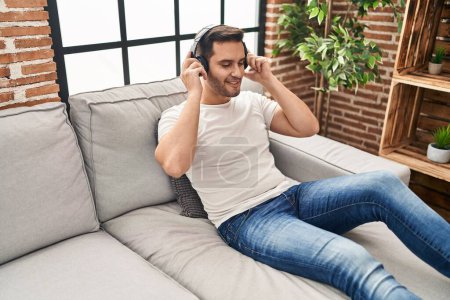 Téléchargez les photos : Jeune homme hispanique écoutant de la musique assis sur le canapé à la maison - en image libre de droit