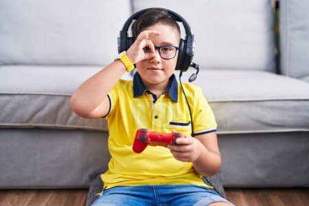 Foto de Young hispanic kid playing video game holding controller wearing headphones doing ok gesture with hand smiling, eye looking through fingers with happy face. - Imagen libre de derechos