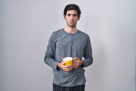 Foto de Young hispanic man wearing pajama drinking a cup of coffee skeptic and nervous, frowning upset because of problem. negative person. - Imagen libre de derechos