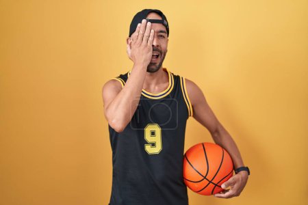 Foto de Middle age bald man holding basketball ball over yellow background covering one eye with hand, confident smile on face and surprise emotion. - Imagen libre de derechos
