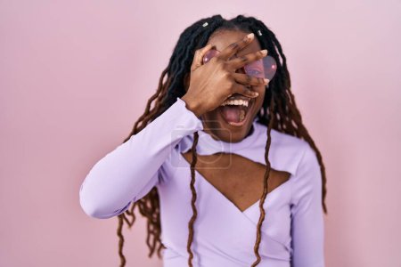 Téléchargez les photos : African woman with braided hair standing over pink background peeking in shock covering face and eyes with hand, looking through fingers with embarrassed expression. - en image libre de droit