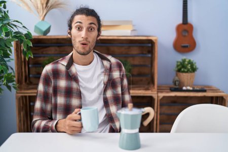 Photo for Young hispanic man drinking coffee from french coffee maker scared and amazed with open mouth for surprise, disbelief face - Royalty Free Image