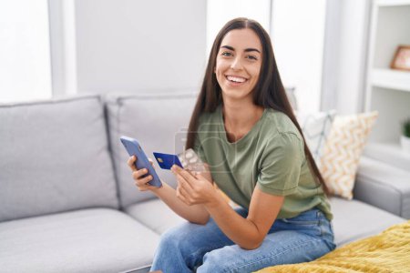 Foto de Joven hermosa mujer hispana usando smartphone y tarjeta de crédito sosteniendo pastillas en casa - Imagen libre de derechos