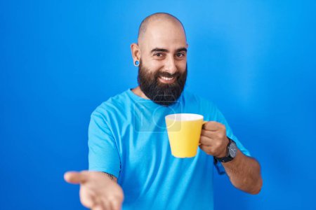 Téléchargez les photos : Jeune homme hispanique avec barbe et tatouages boire une tasse de café souriant gai offrant main de palme donnant assistance et acceptation. - en image libre de droit