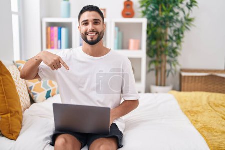 Téléchargez les photos : Jeune homme hispanique utilisant ordinateur portable assis sur le lit souriant heureux pointant avec la main et le doigt - en image libre de droit