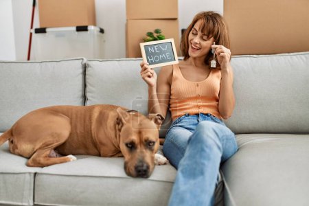 Photo for Young caucasian woman holding key and new home blackboard sitting on sofa with dog at home - Royalty Free Image