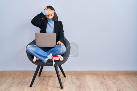 Téléchargez les photos : Jeune femme hispanique assise sur une chaise en utilisant un ordinateur portable souriant et riant avec la main sur le visage couvrant les yeux pour la surprise. concept aveugle. - en image libre de droit