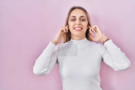 Photo for Young blonde woman wearing white sweater over pink background covering ears with fingers with annoyed expression for the noise of loud music. deaf concept. - Royalty Free Image