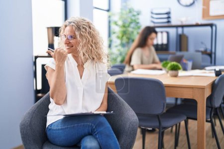 Téléchargez les photos : Femme d'âge moyen travaillant au bureau tenant presse-papiers pointant pouce vers le côté souriant heureux avec bouche ouverte - en image libre de droit