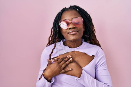 Téléchargez les photos : Femme africaine avec les cheveux tressés debout sur fond rose souriant avec les mains sur la poitrine avec les yeux fermés et geste reconnaissant sur le visage. concept de santé. - en image libre de droit