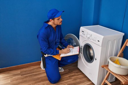 Photo for Young hispanic man technician repairing washing machine writing on document at laundry room - Royalty Free Image