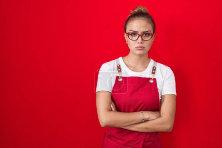 Photo for Young hispanic woman wearing waitress apron over red background skeptic and nervous, disapproving expression on face with crossed arms. negative person. - Royalty Free Image