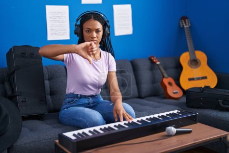 Foto de African american woman with braids playing piano keyboard at music studio with angry face, negative sign showing dislike with thumbs down, rejection concept - Imagen libre de derechos