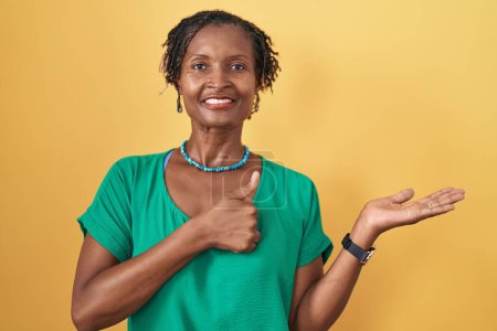 Téléchargez les photos : African woman with dreadlocks standing over yellow background showing palm hand and doing ok gesture with thumbs up, smiling happy and cheerful - en image libre de droit
