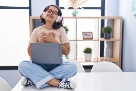 Photo for Young hispanic woman using laptop sitting on the table wearing headphones smiling with hands on chest with closed eyes and grateful gesture on face. health concept. - Royalty Free Image