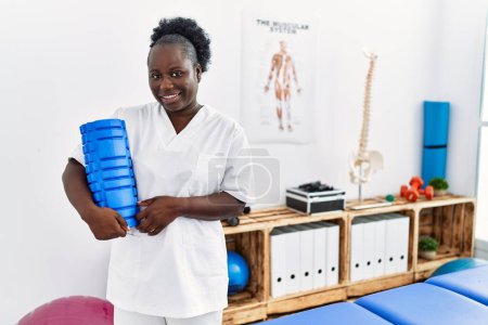 Foto de Mujer afroamericana joven con uniforme de fisioterapeuta sosteniendo rodillo de espuma en la clínica - Imagen libre de derechos