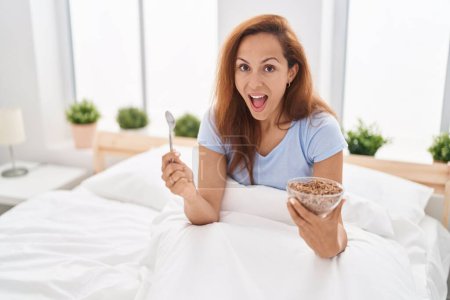 Téléchargez les photos : Brunette femme manger le petit déjeuner dans le lit célébrant fou et étonné pour le succès avec les yeux ouverts hurlant excité. - en image libre de droit