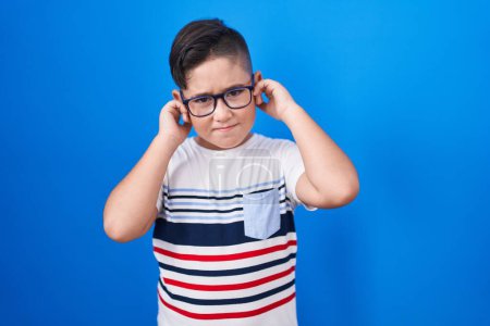 Photo for Young hispanic kid standing over blue background covering ears with fingers with annoyed expression for the noise of loud music. deaf concept. - Royalty Free Image