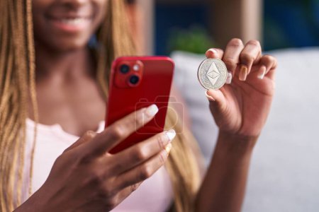 Téléchargez les photos : African american woman holding ethereum classic crypto currency using smartphone at home - en image libre de droit