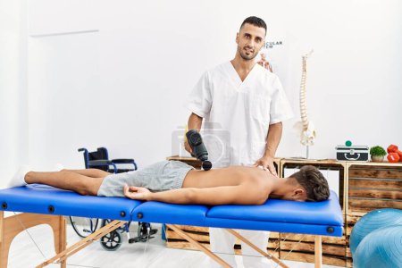 Foto de Dos hombres hispanos fisioterapeuta y paciente masajeando la espalda usando pistola de percusión en la clínica - Imagen libre de derechos