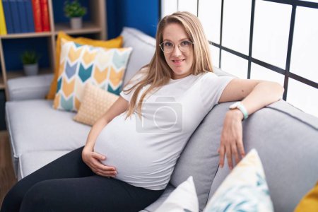 Foto de Young pregnant woman touching belly sitting on sofa at home - Imagen libre de derechos