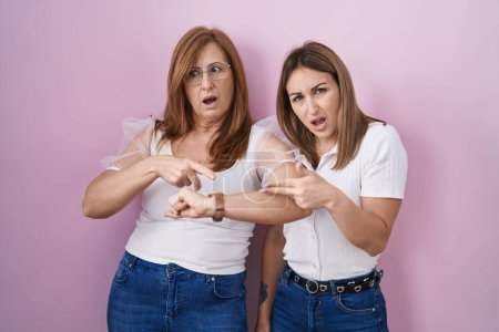 Téléchargez les photos : Mère et fille hispaniques portant un t-shirt blanc décontracté sur fond rose à la hâte pointant vers regarder le temps, l'impatience, contrarié et en colère pour retard de délai - en image libre de droit