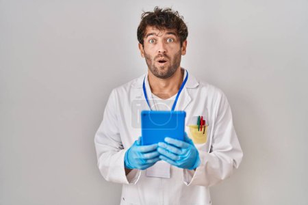 Foto de Hombre científico hispano trabajando con tableta asustado y sorprendido con la boca abierta para sorpresa, cara de incredulidad - Imagen libre de derechos