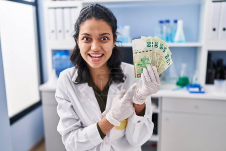 Téléchargez les photos : Hispanic woman with dark hair working at scientist laboratory holding money smiling happy pointing with hand and finger - en image libre de droit