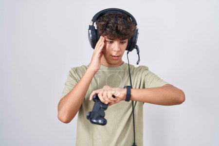 Téléchargez les photos : Hispanic teenager playing video game holding controller looking at the watch time worried, afraid of getting late - en image libre de droit