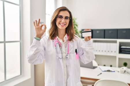 Foto de Joven doctora sosteniendo tarjeta de crédito en la clínica haciendo ok signo con los dedos, sonriendo gesto amistoso excelente símbolo - Imagen libre de derechos