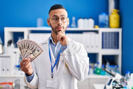Téléchargez les photos : Young hispanic man working at scientist laboratory holding money serious face thinking about question with hand on chin, thoughtful about confusing idea - en image libre de droit