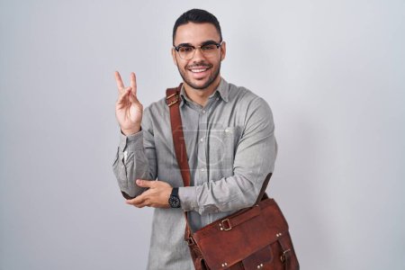 Téléchargez les photos : Jeune homme hispanique portant une valise souriant avec un clin d'œil heureux à la caméra faisant signe de victoire avec les doigts. numéro deux. - en image libre de droit
