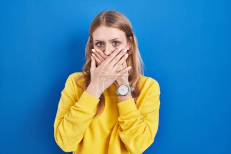 Photo for Young caucasian woman standing over blue background shocked covering mouth with hands for mistake. secret concept. - Royalty Free Image