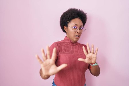 Téléchargez les photos : Beautiful african woman with curly hair standing over pink background afraid and terrified with fear expression stop gesture with hands, shouting in shock. panic concept. - en image libre de droit