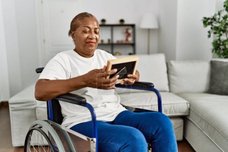 Foto de Mujer afroamericana mayor mirando foto sentada en silla de ruedas en casa - Imagen libre de derechos