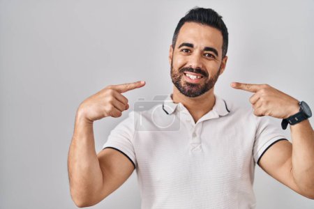 Téléchargez les photos : Jeune homme hispanique avec barbe portant des vêtements décontractés sur fond blanc souriant et joyeux montrant et pointant avec les doigts les dents et la bouche. concept de santé dentaire. - en image libre de droit