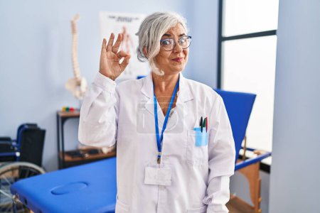 Photo for Middle age woman with grey hair working at pain recovery clinic smiling positive doing ok sign with hand and fingers. successful expression. - Royalty Free Image