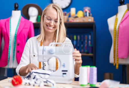 Foto de Joven mujer rubia sastre sonriendo confiado usando máquina de coser en el estudio de costura - Imagen libre de derechos