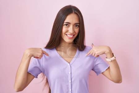 Foto de Mujer hispana joven con el pelo largo de pie sobre fondo rosa mirando confiado con sonrisa en la cara, señalándose con los dedos orgullosos y felices. - Imagen libre de derechos