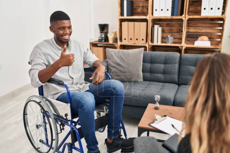 Photo for African american man doing therapy sitting on wheelchair doing happy thumbs up gesture with hand. approving expression looking at the camera showing success. - Royalty Free Image