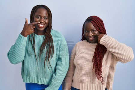 Foto de Dos mujeres africanas de pie sobre fondo azul señalando con el dedo de la mano a la cara y la nariz, sonriendo alegre. concepto de belleza - Imagen libre de derechos