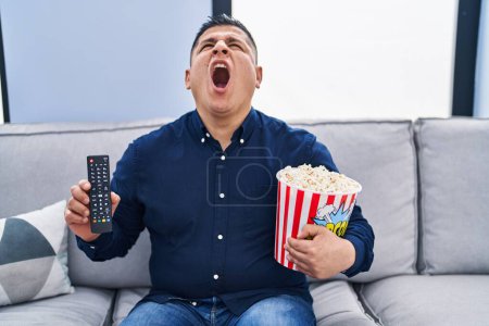 Photo for Hispanic young man eating popcorn using tv control angry and mad screaming frustrated and furious, shouting with anger looking up. - Royalty Free Image
