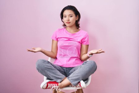 Photo for Hispanic young woman sitting on chair over pink background clueless and confused with open arms, no idea concept. - Royalty Free Image