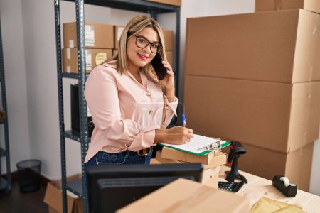 Foto de Young hispanic woman ecommerce business worker talking on the smartphone and writing on clipboard at office - Imagen libre de derechos