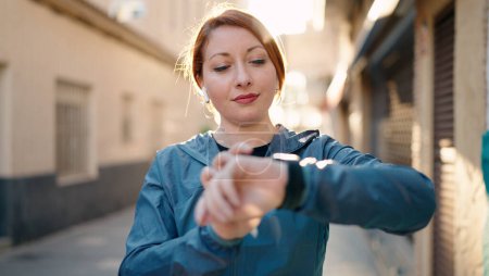Téléchargez les photos : Young redhead woman wearing sportswear listening to music looking stopwatch at street - en image libre de droit