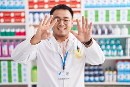 Téléchargez les photos : Jeune homme chinois travaillant à la pharmacie pharmacie montrant et pointant vers le haut avec les doigts numéro neuf tout en souriant confiant et heureux. - en image libre de droit