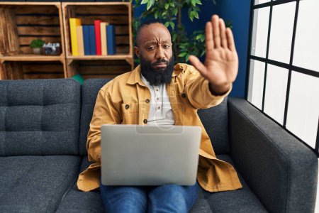 Foto de African american man using laptop at home sitting on the sofa with open hand doing stop sign with serious and confident expression, defense gesture - Imagen libre de derechos