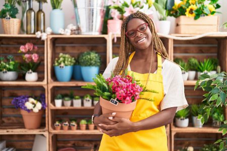 Téléchargez les photos : Femme afro-américaine avec les cheveux tressés travaillant à la boutique de fleuriste tenant l'usine regardant positif et heureux debout et souriant avec un sourire confiant montrant des dents - en image libre de droit